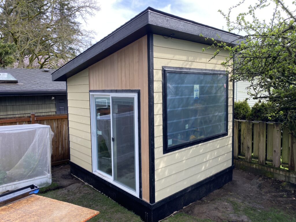 A small cream-colored shed with a slanted roof, large side window, and sliding glass door stands in a backyard near a wooden fence and greenery.
