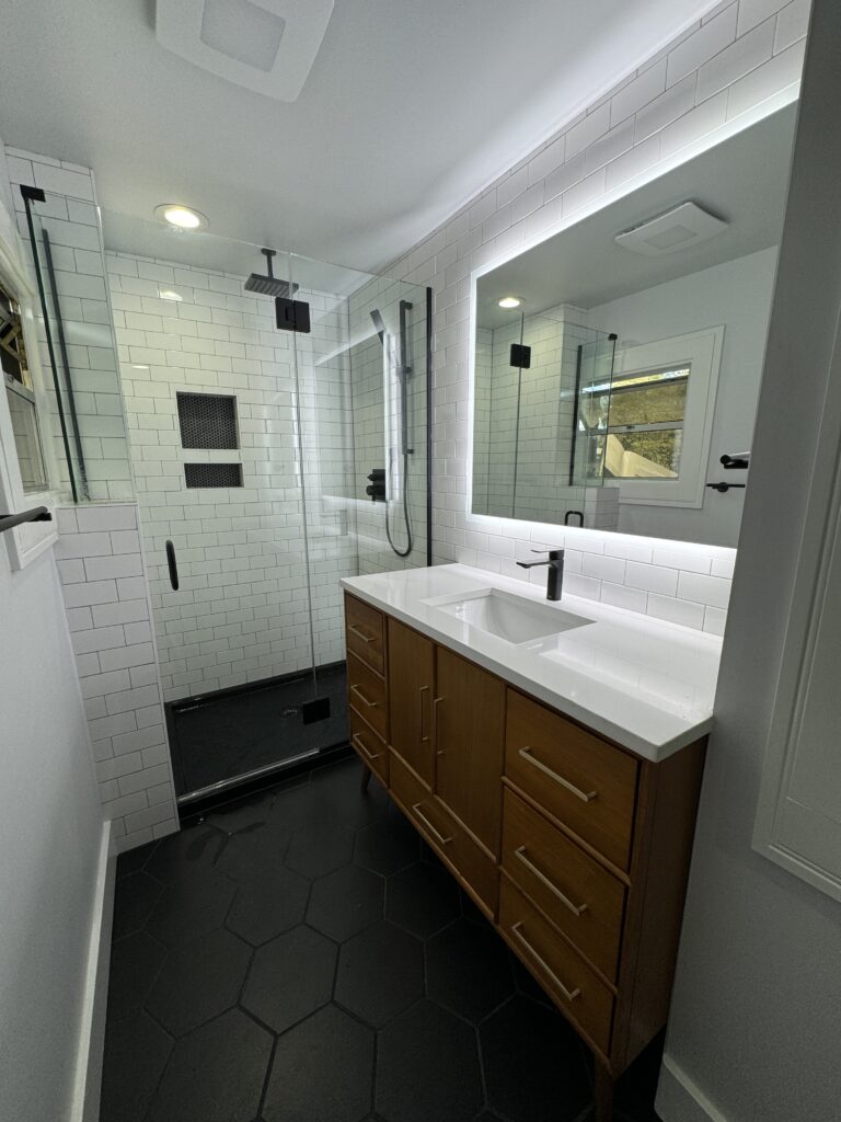 Modern bathroom with black hexagon tile floor, glass shower with white subway tiles, a wooden vanity with double sinks, and a large backlit mirror above the sink.