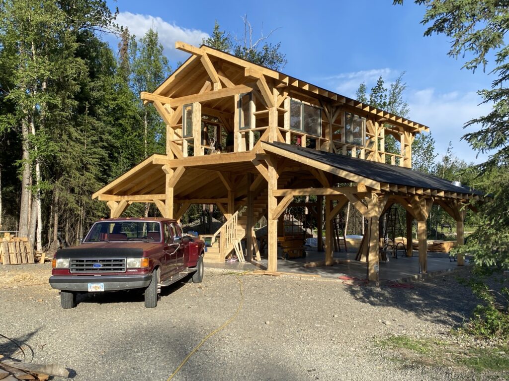 A wooden, two-story cabin with large windows and an open ground floor stands in a forest clearing. A red pickup truck is parked in front, and sunlight filters through the trees and onto the gravel driveway.