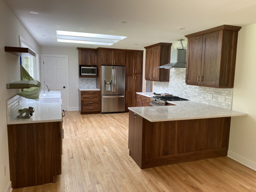 Modern kitchen with wooden cabinets, stainless steel appliances, white countertops, a farmhouse sink, and light wood flooring. There is a skylight above, and the backsplash is made of white tiles.