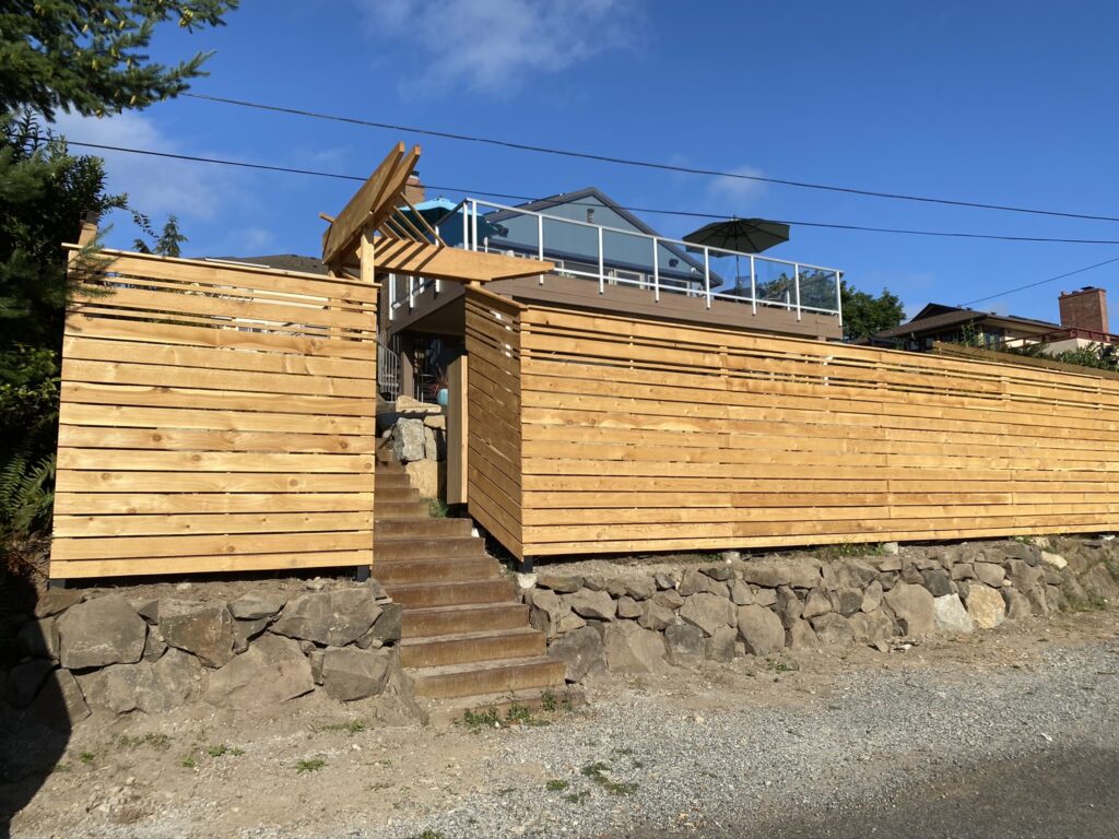 A modern house with a glass balcony sits behind a tall wooden privacy fence and gate, with stone steps leading up from a gravel road. The sky is clear and blue.