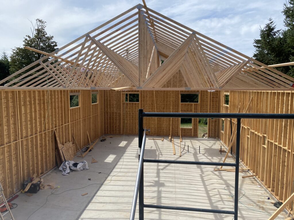 A wooden house frame under construction, showing exposed wall studs and roof trusses, with tools and materials scattered on the concrete foundation and a metal scaffold in the foreground.