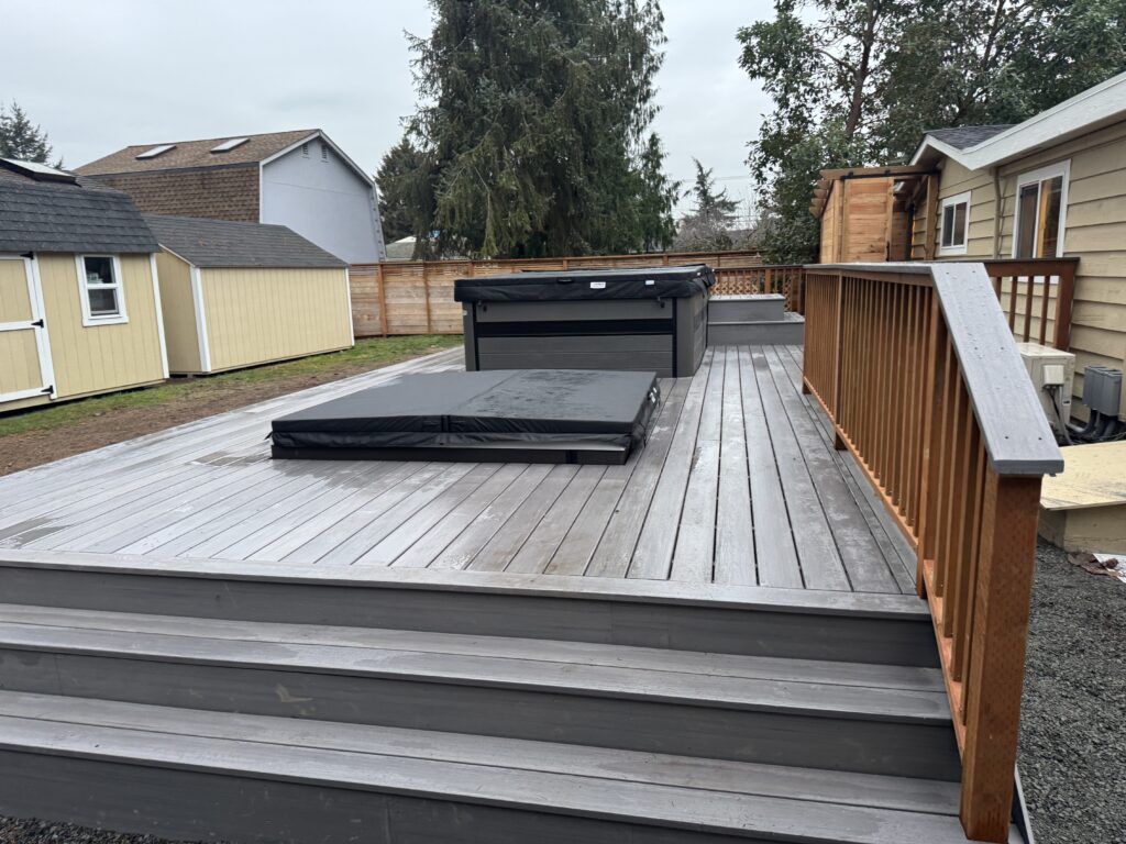 A wooden backyard deck with steps, featuring a covered hot tub and a covered pool. The deck is bordered by a wooden railing, and there are sheds, trees, and a house visible in the background under a cloudy sky.
