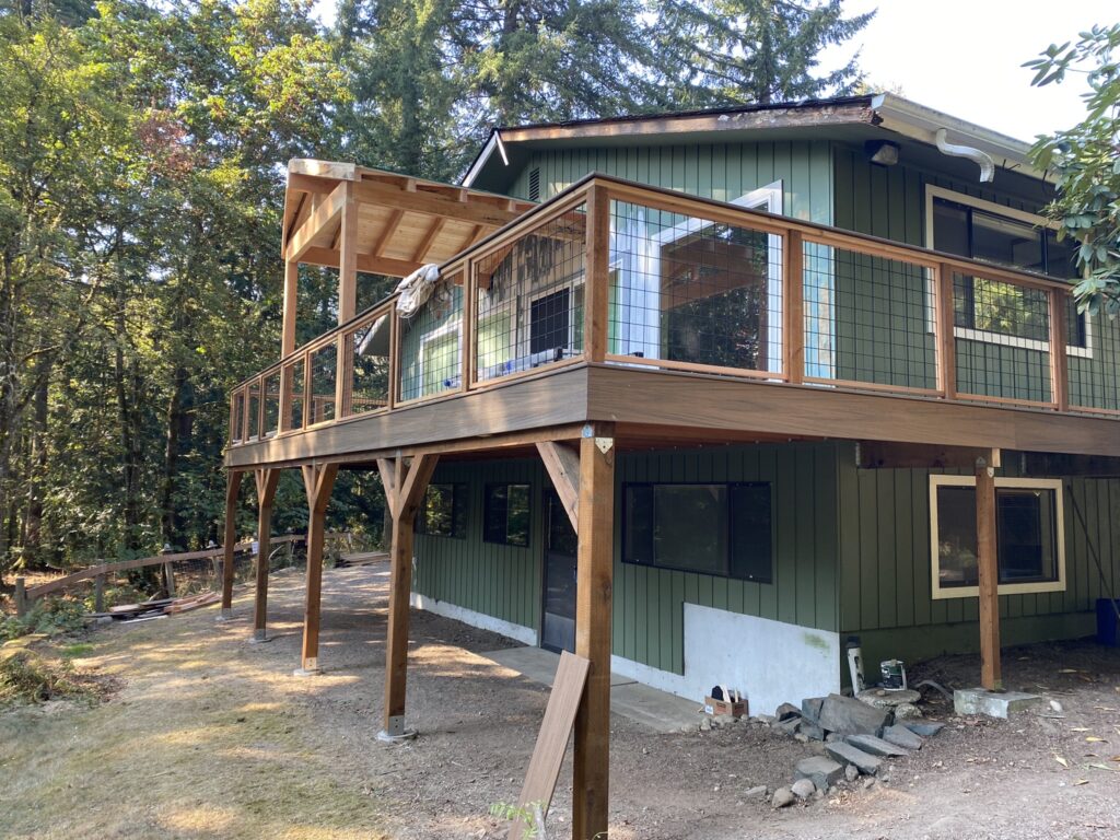 A green two-story house with a large wooden deck and metal railings, surrounded by trees and sunlight. The deck is elevated on wooden posts, and there are building materials on the ground below.