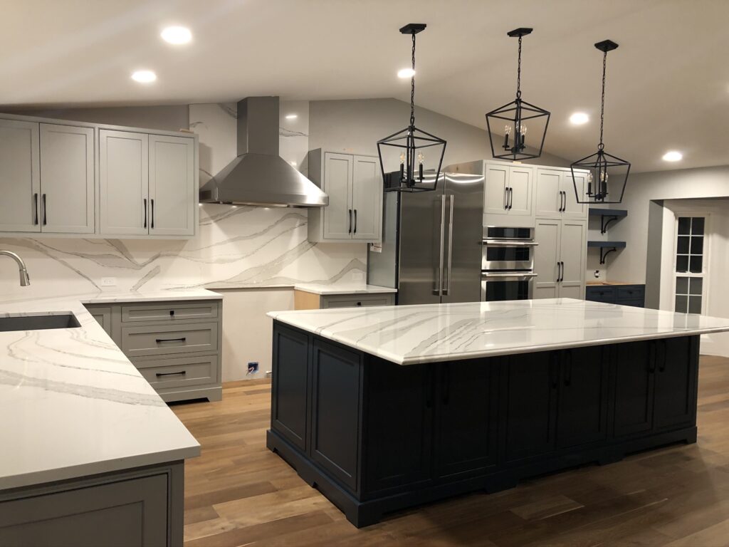 Modern kitchen with gray cabinets, stainless steel appliances, and a large white marble island. Three black pendant lights hang above the island, and wood flooring adds warmth to the space.