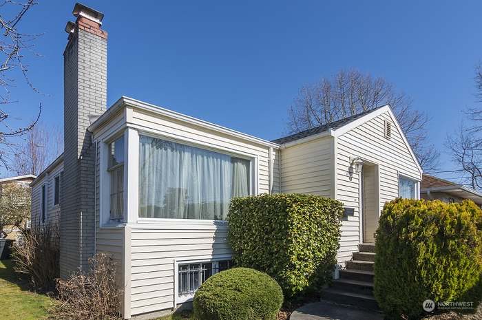 Single-story beige house with large front window, gray chimney, and neatly trimmed bushes along the walkway. The sky is clear and blue, and bare trees are visible in the background.