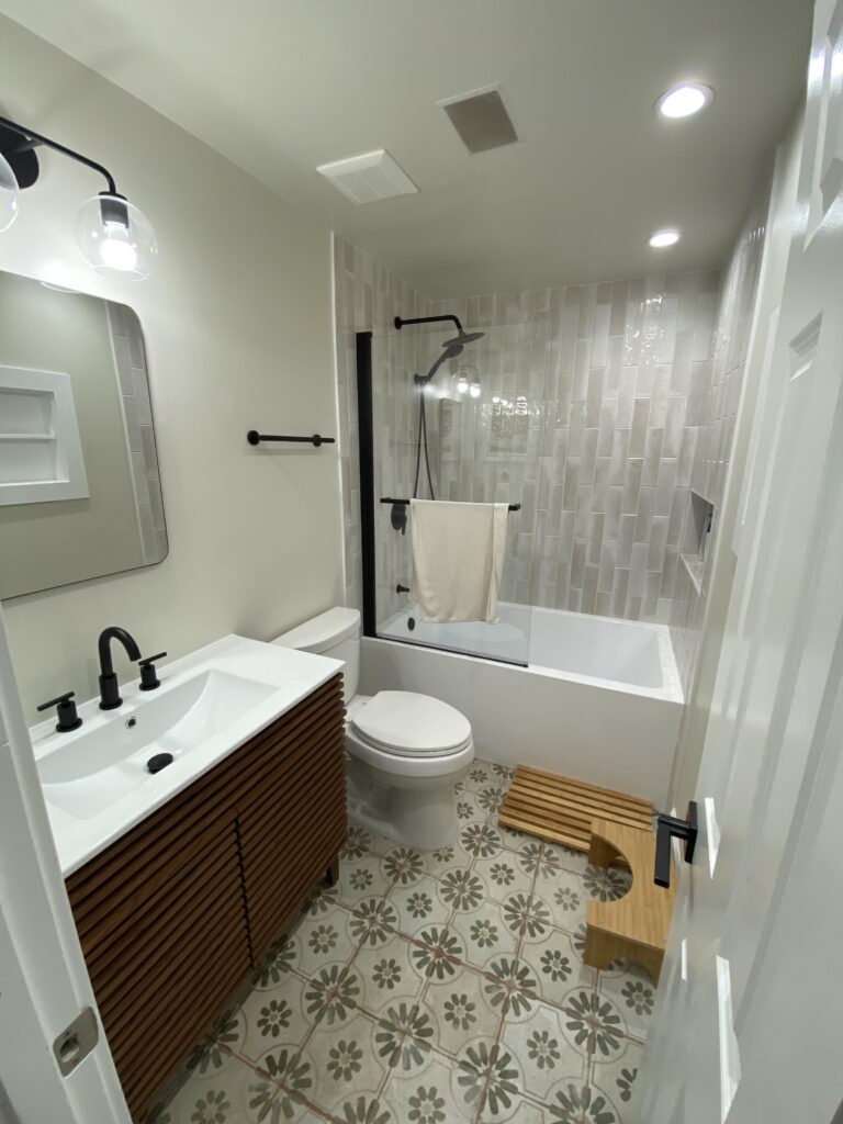Modern bathroom with patterned tile floor, wooden vanity, white sink, wall-mounted mirror, toilet, bathtub with glass partition, white towel on black rod, and a wooden step stool on the floor. Bright overhead lighting.