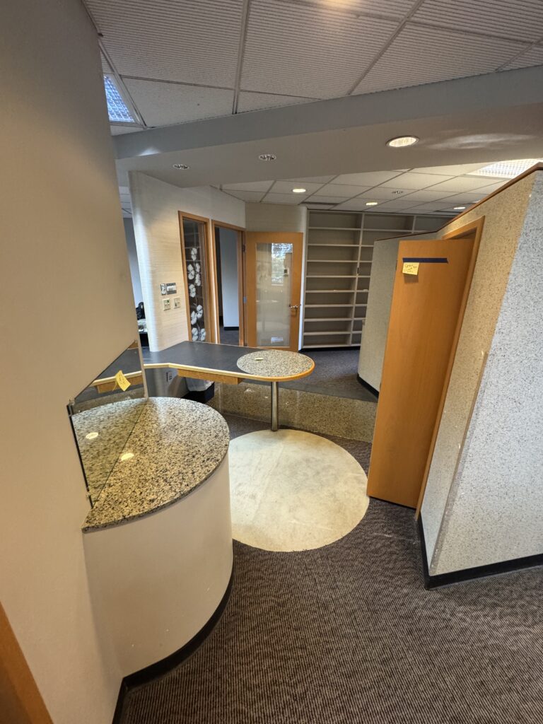 A small office area with granite countertops, wood-paneled surfaces, shelves, and open doorways. The space has a circular desk and modern design elements, with neutral colors and office lighting.