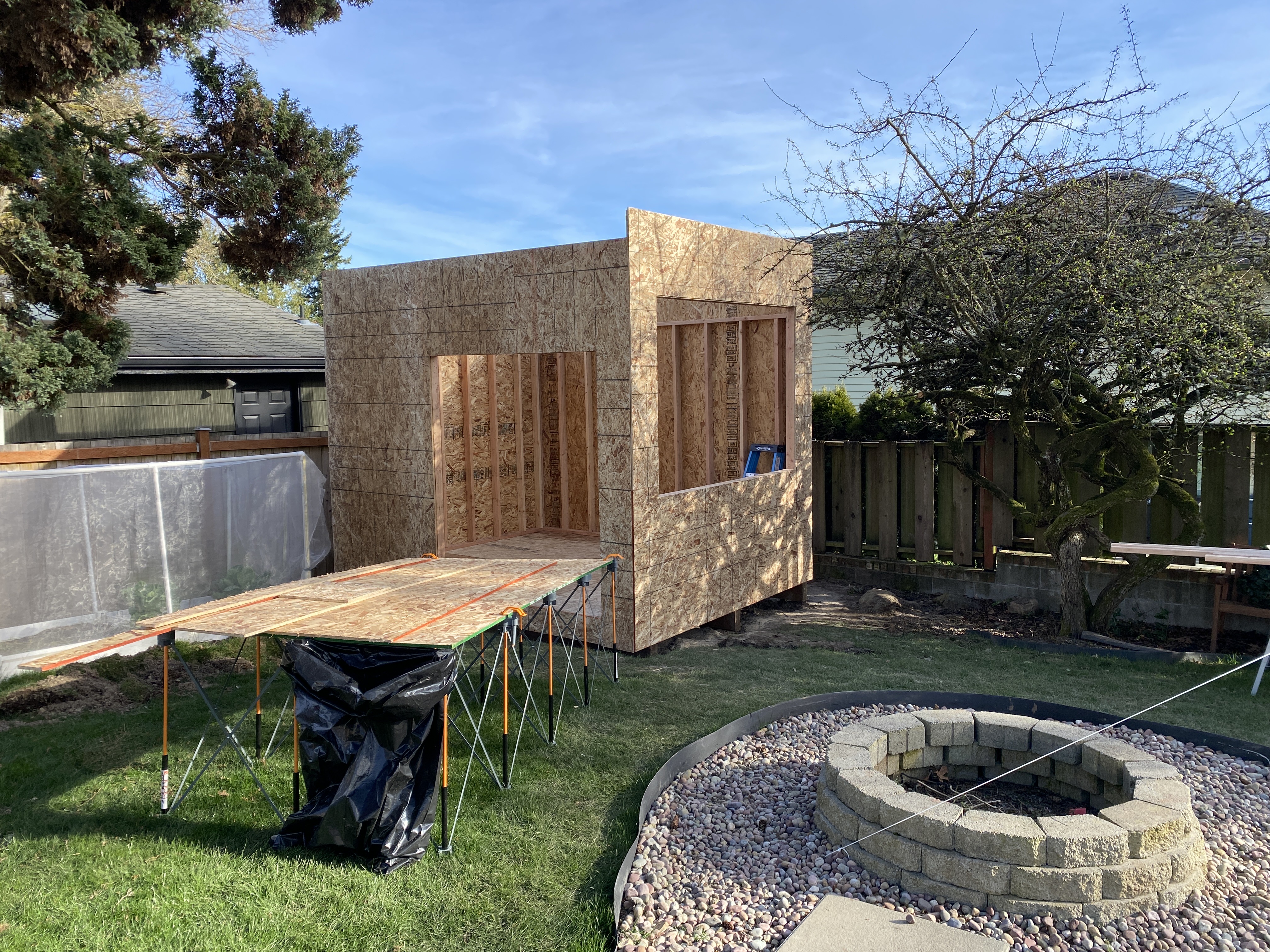 Detached studio framed and sheathed with door and large window openings cut, beside a backyard fire pit in Bothell
