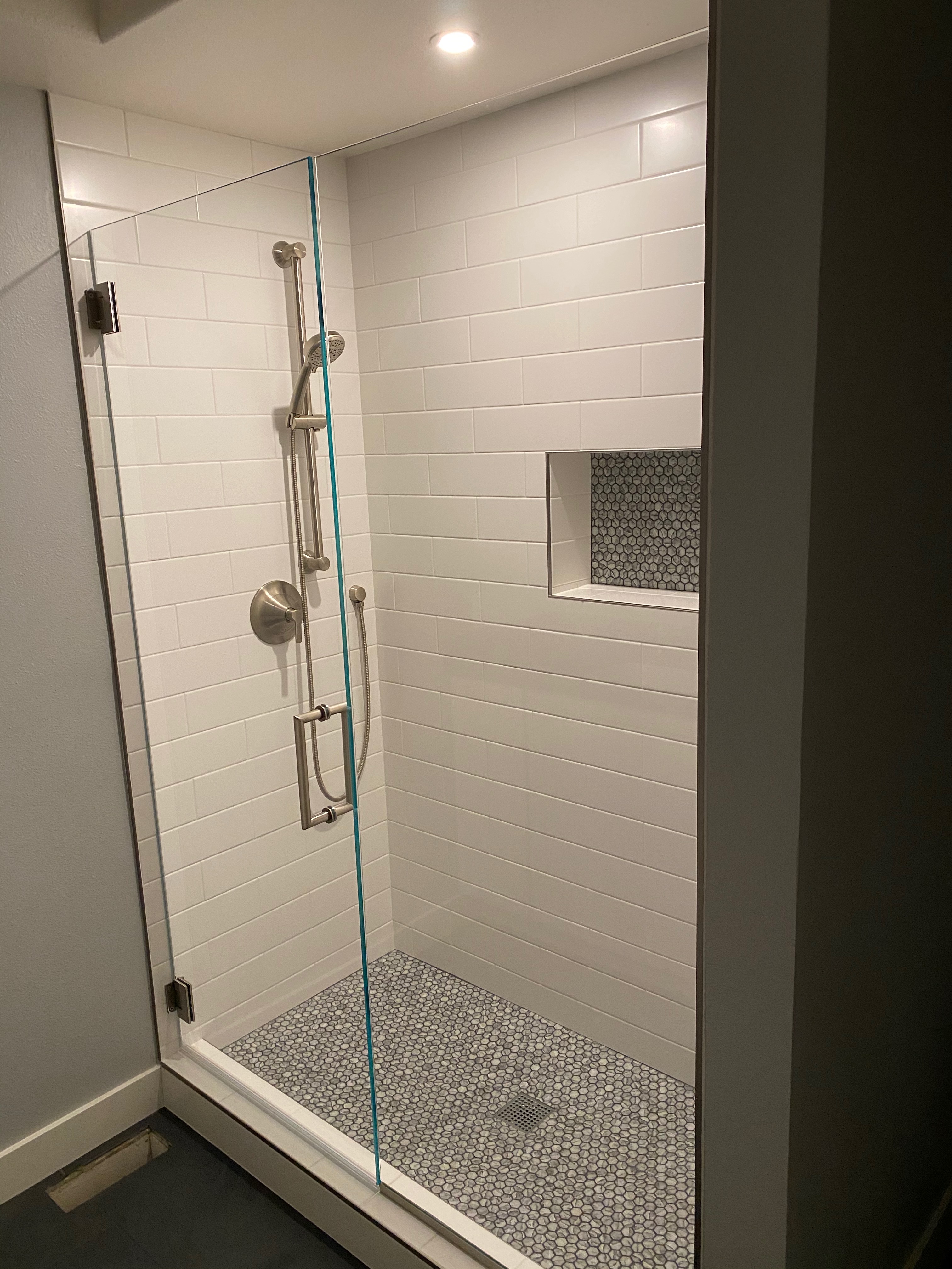 Remodeled bathroom shower with white subway tile, a recessed pebble-tile niche, pebble shower floor, frameless glass panel, and brushed nickel handheld shower on a slide bar