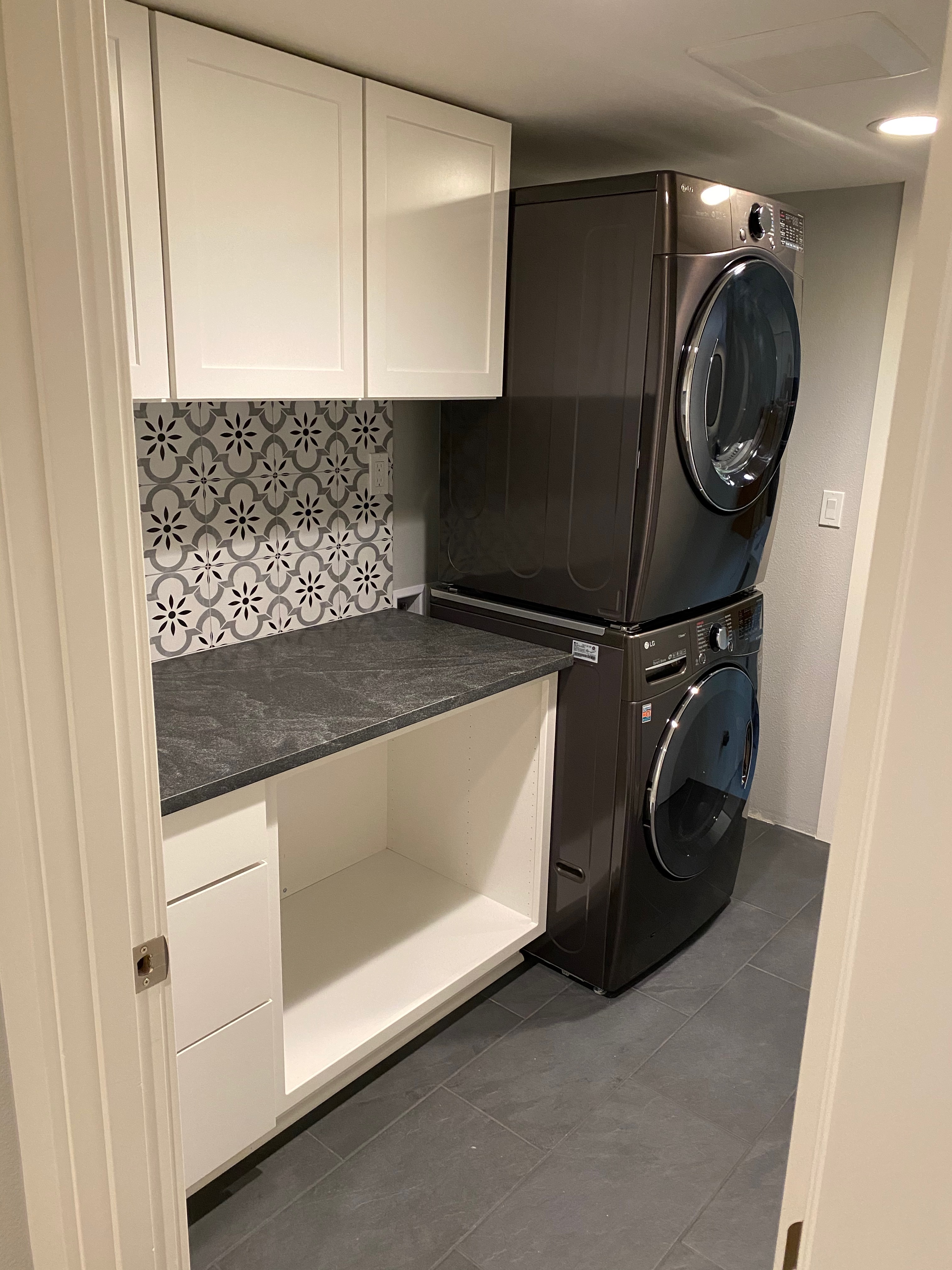 Custom laundry room after remodel with white shaker cabinetry, dark stone countertop, black and white patterned tile backsplash, slate tile floor, and a stacked LG washer and dryer