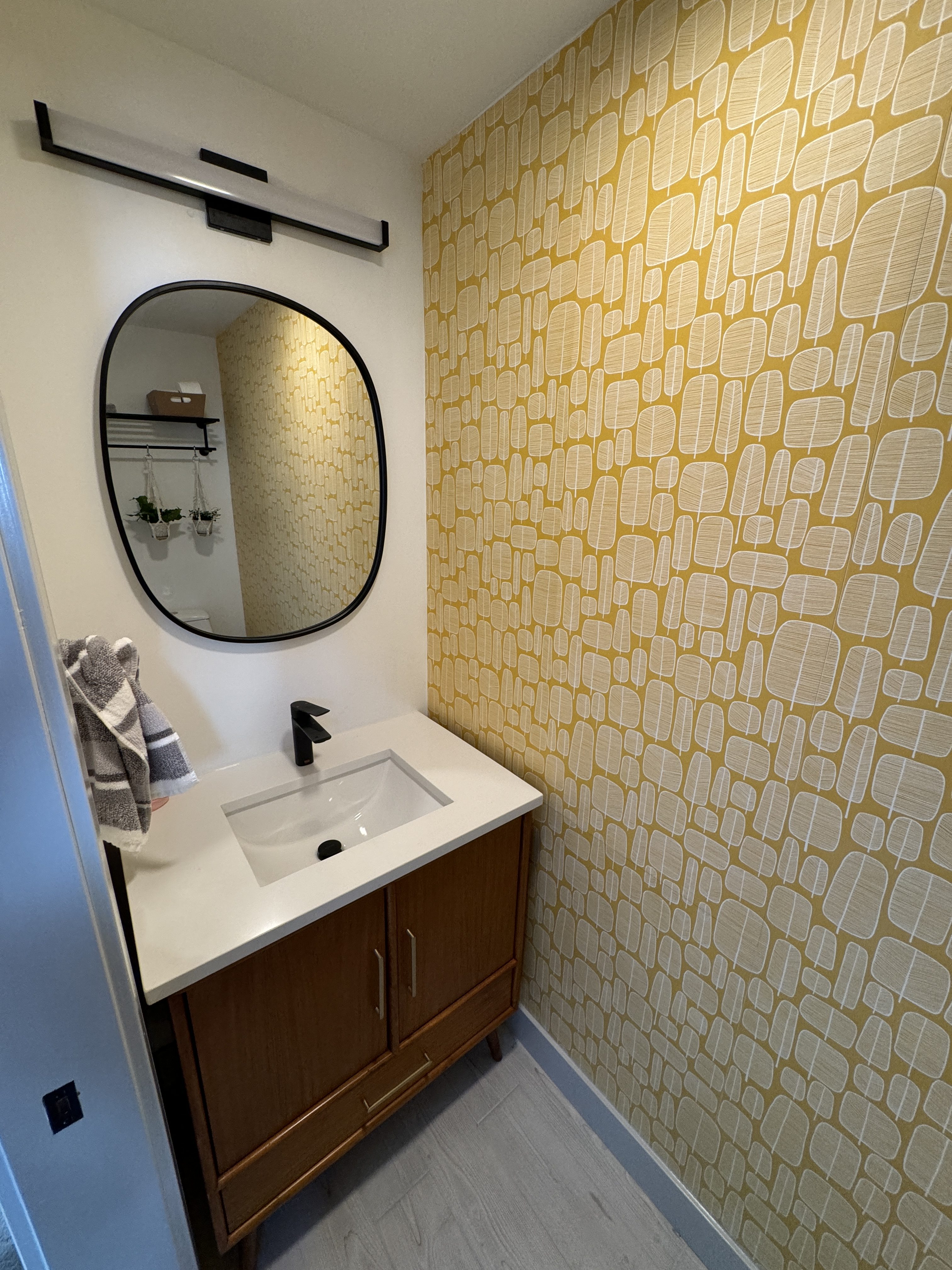 New Bellevue guest powder room with ochre patterned wallpaper, an oval mirror, and a walnut vanity with a black faucet