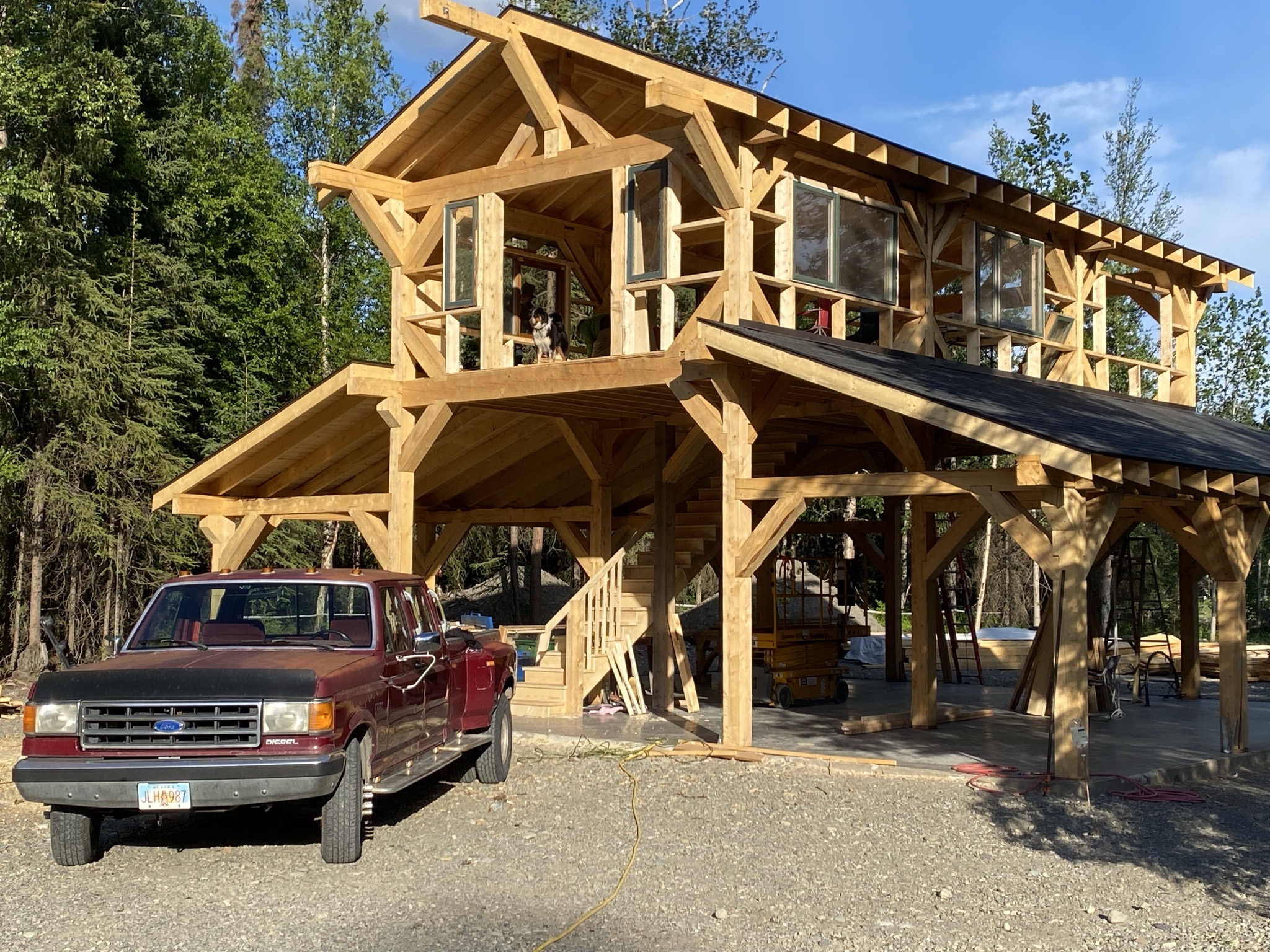 Standing timber frame barndominium in daylight with a dog on the loft and a red pickup truck