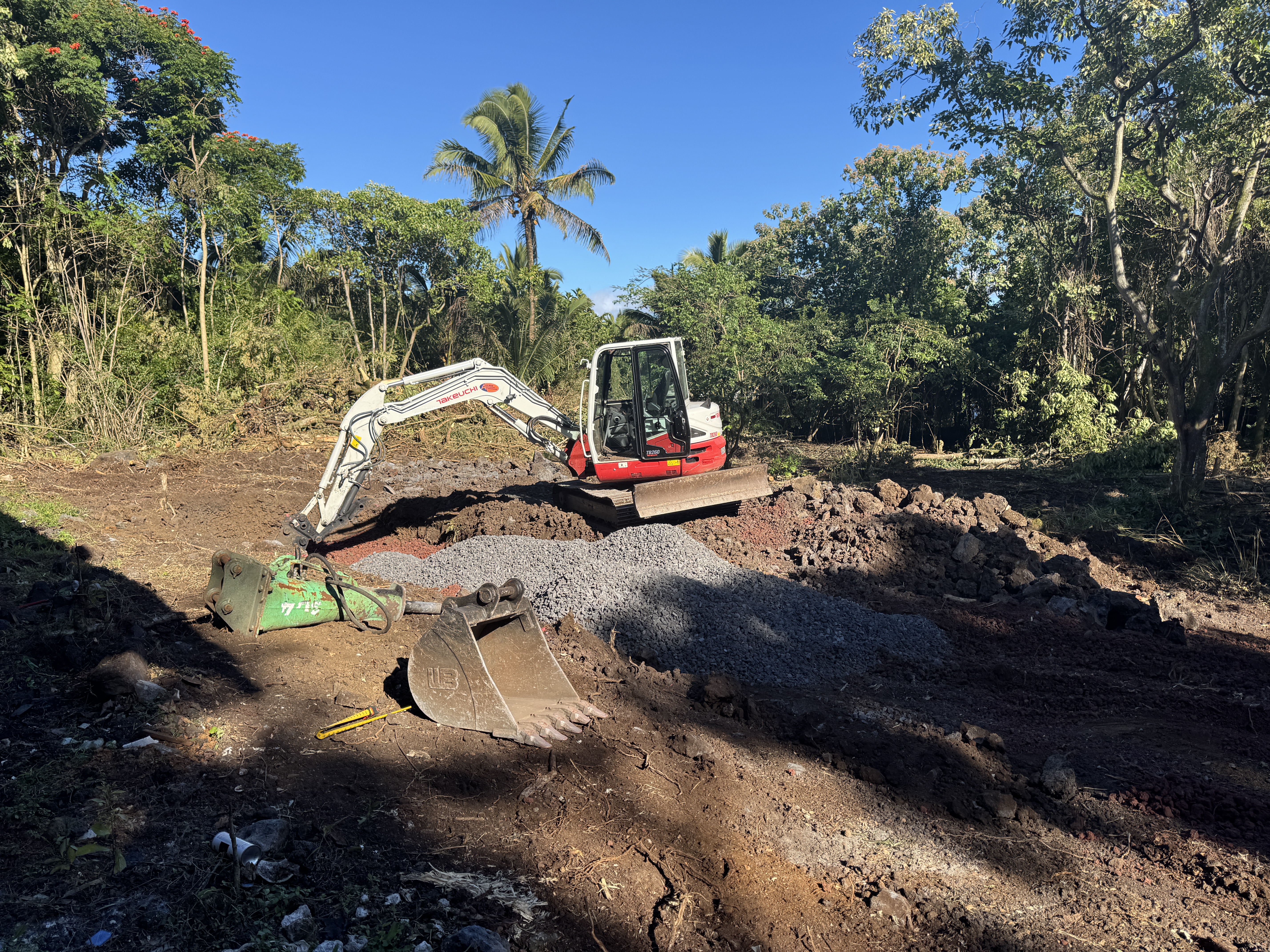A compact excavator clearing dense jungle and grading a pad on lava rock at the start of the homestead build