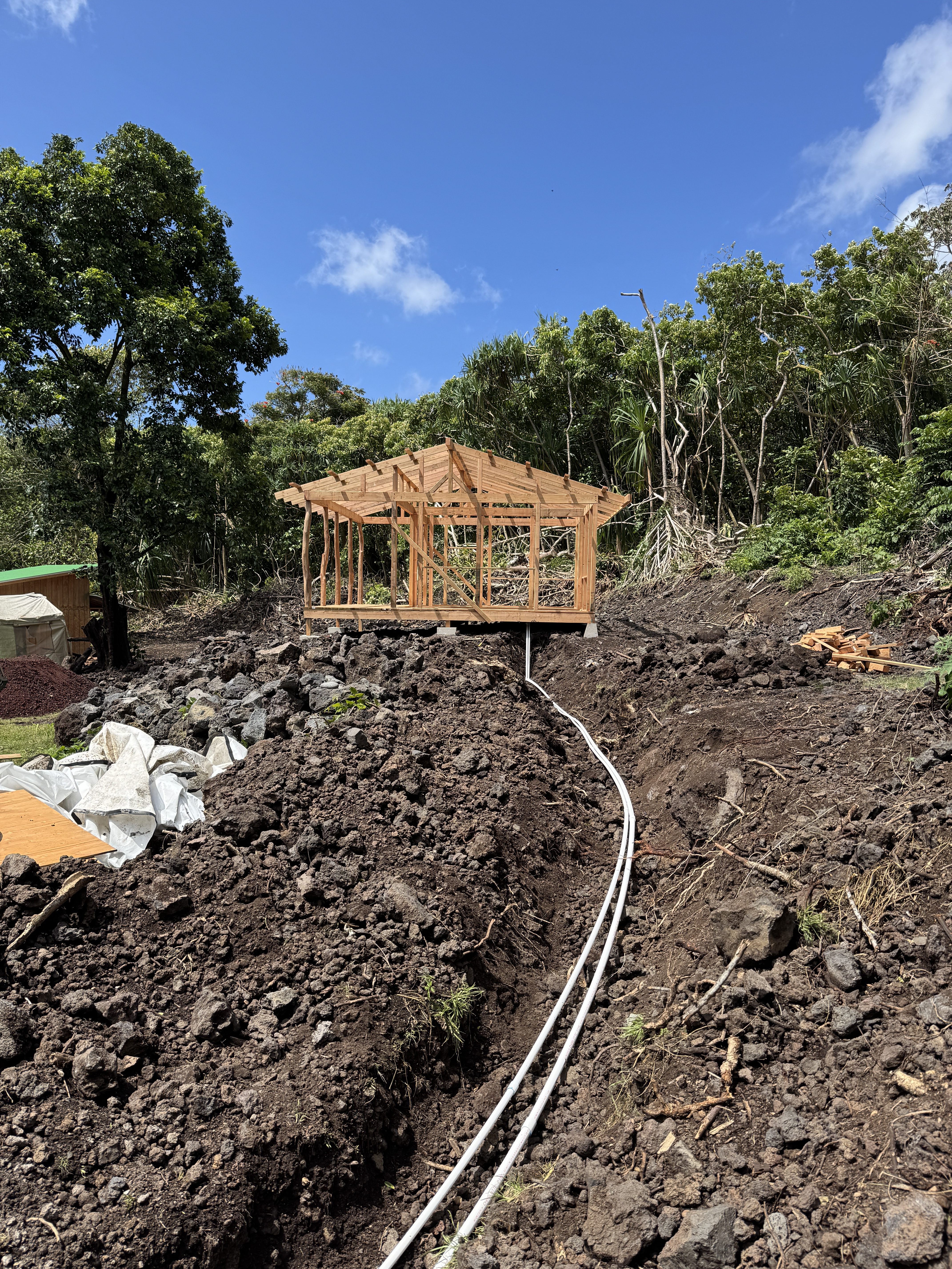 Water and utility lines running through an open trench in lava rock toward a newly framed structure